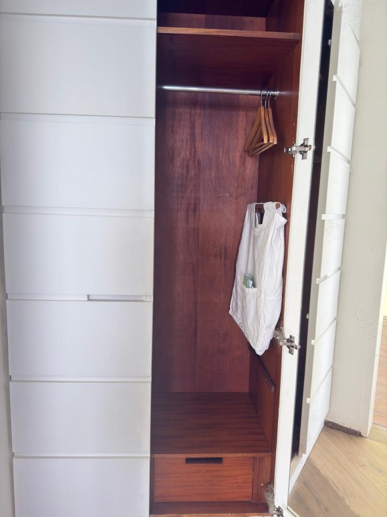 A wooden closet with an open door, showing a few wooden hangers on a metal rod and a white reusable bag hanging inside. There is also a small drawer at the bottom of the closet.