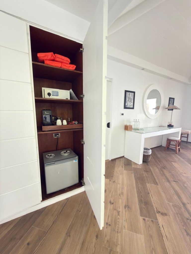 A closet with shelves holding life jackets, a safe, a kettle, cups, and a mini fridge; the closet opens into a modern room with wood floors, a console table, and a circular mirror on the wall.
