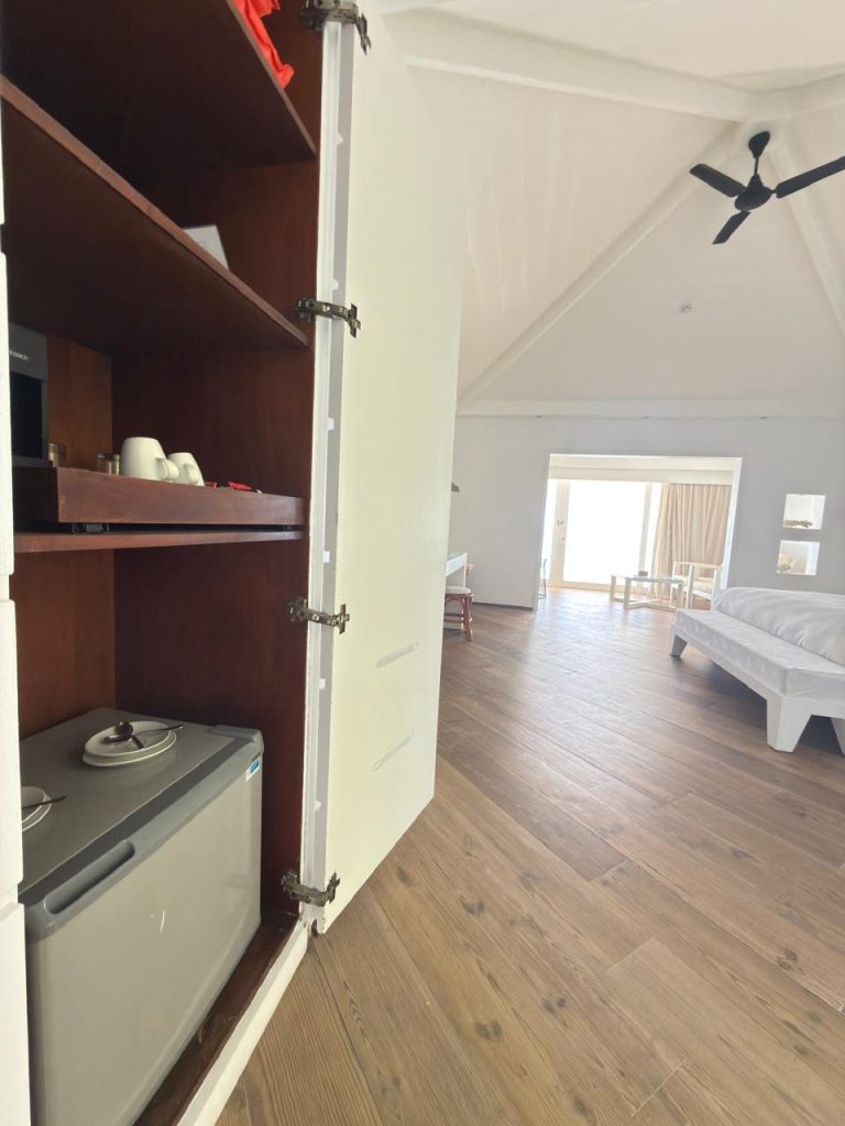 A hotel room with wooden floors, showing an open cabinet containing a mini fridge and tea set on shelves. In the background, a bed, desk, and large windows letting in sunlight are visible.