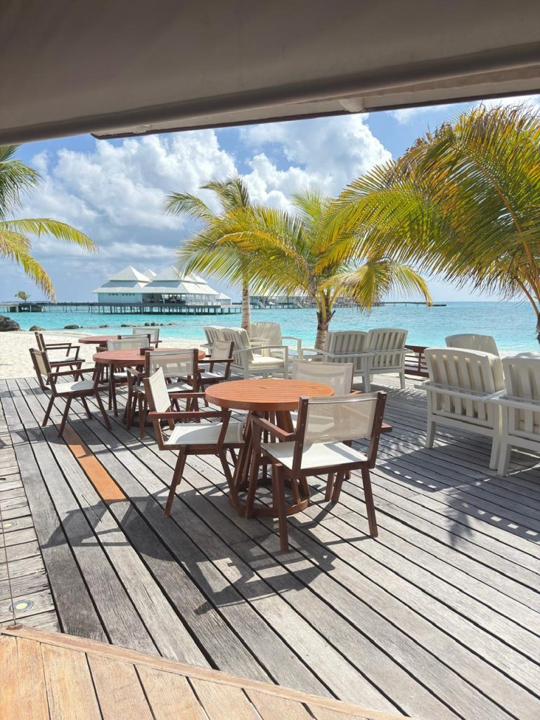Outdoor beachside dining area with wooden tables and chairs on a wooden deck, surrounded by palm trees. The turquoise ocean and a pavilion on stilts are visible in the background under a partly cloudy sky.