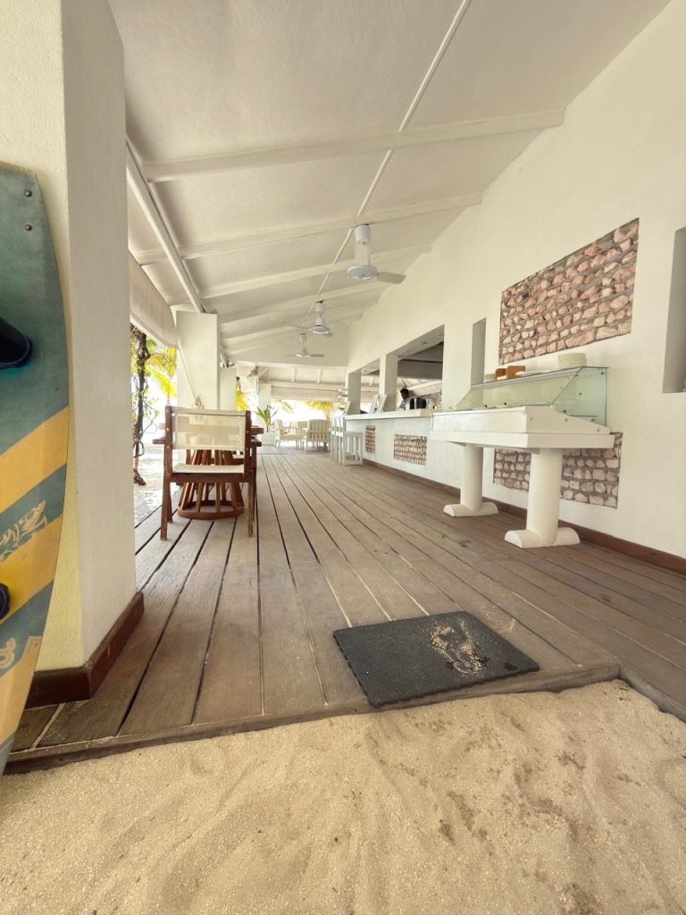 A beachside café with wooden floors, white walls, a serving counter, ceiling fans, and chairs and tables. Sand covers part of the floor, with a surfboard leaning against the wall on the left. Palm trees visible outside.