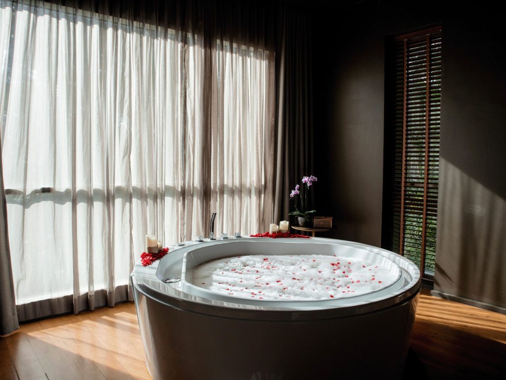 A round, white bathtub filled with bubble bath and rose petals sits by large windows with sheer curtains. Candles, a potted orchid, and spa items are arranged around the tub, creating a relaxing, serene atmosphere.