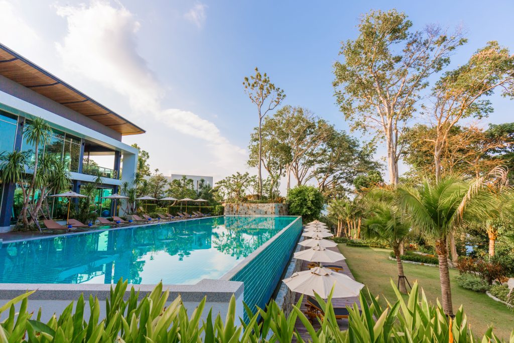 A modern building with large windows overlooks a tranquil infinity pool surrounded by lounge chairs and umbrellas, lush greenery, and tall trees under a blue sky with scattered clouds.