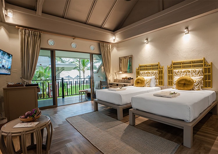 A cozy hotel room with two single beds, yellow headboards, decorative pillows, a desk, TV, and sliding glass doors opening to a balcony with a view of palm trees and outdoor loungers. Warm lighting and wooden decor create a relaxing atmosphere.