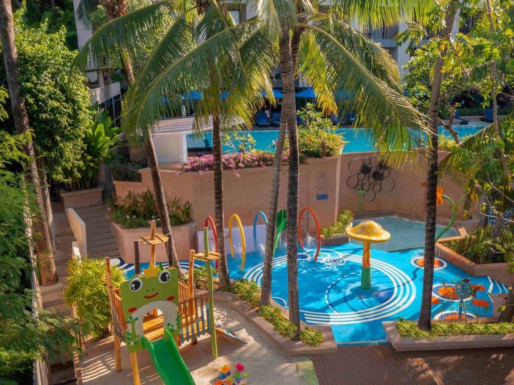 Children’s playground and splash pad under palm trees, featuring a green frog slide, colorful climbing structures, water sprayers, and a mushroom fountain, with a swimming pool and lush greenery in the background.