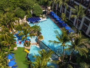 Aerial view of a resort pool surrounded by palm trees, lounge chairs with blue umbrellas, and a green lawn area. The pool has a unique shape, small waterfalls, and is bordered by a multi-story building.