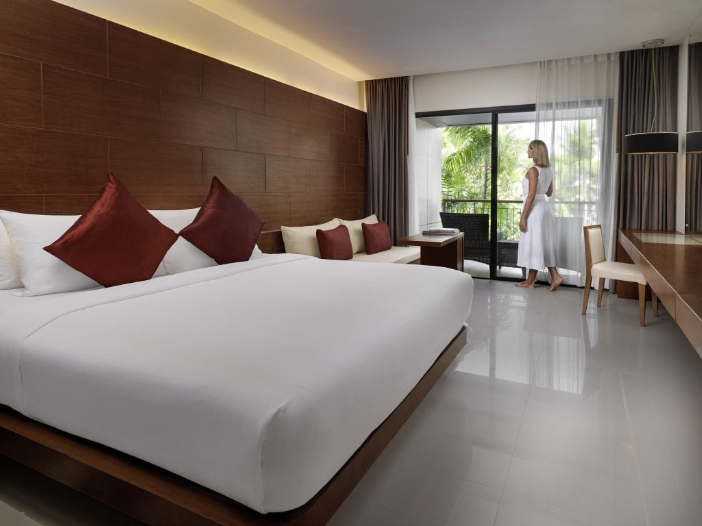 Modern hotel room with a large bed, white linens, and red pillows. A woman in a white dress stands by the window looking outside. The room features wood paneling, a sofa, desk, and a balcony with greenery visible.