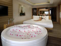 Spacious hotel room with a large circular bathtub filled with water and pink flower petals in the foreground, and a person relaxing on a neatly made bed in the background.