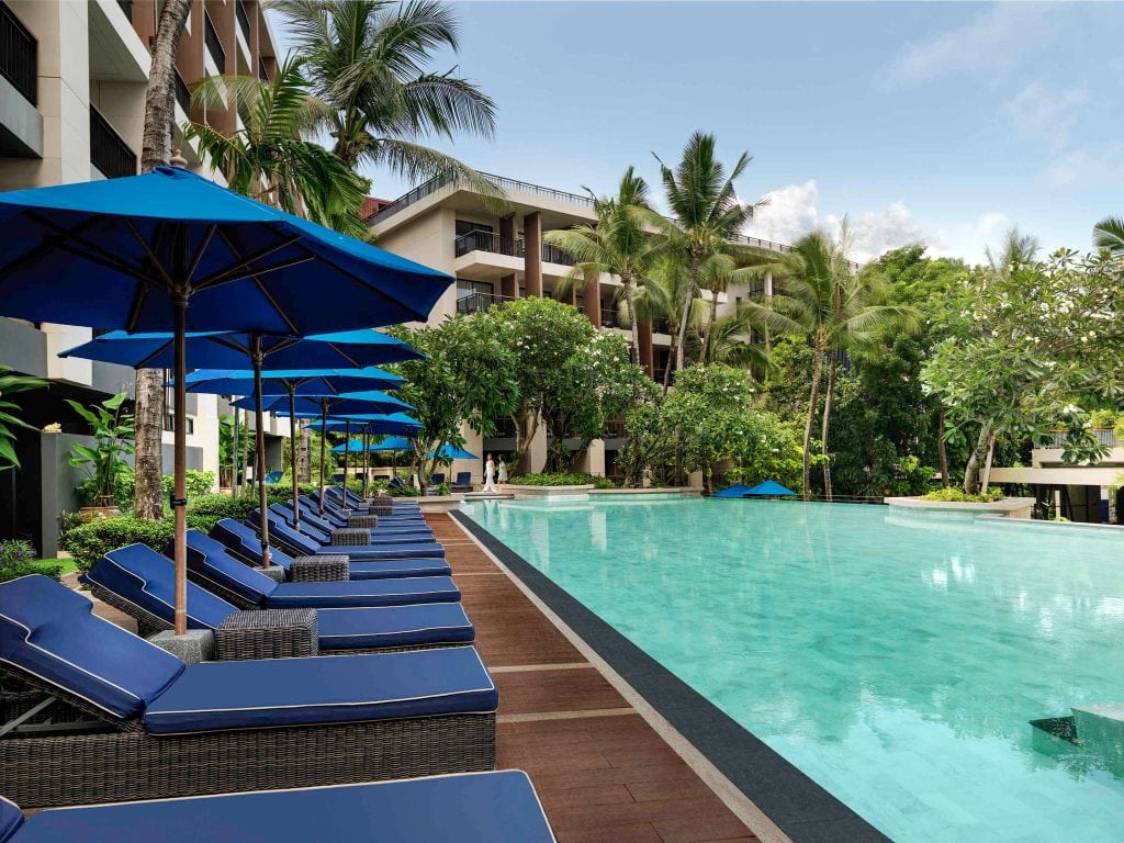 A luxury hotel poolside with blue lounge chairs and umbrellas, surrounded by palm trees and lush greenery, with modern buildings in the background under a clear sky.