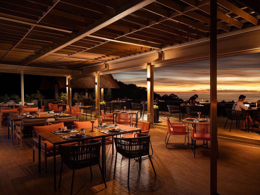 A stylish, open-air restaurant with neatly set tables and orange chairs overlooks a scenic sunset, casting a warm glow over the space. Few diners enjoy the view and ambiance in a relaxed, elegant atmosphere.