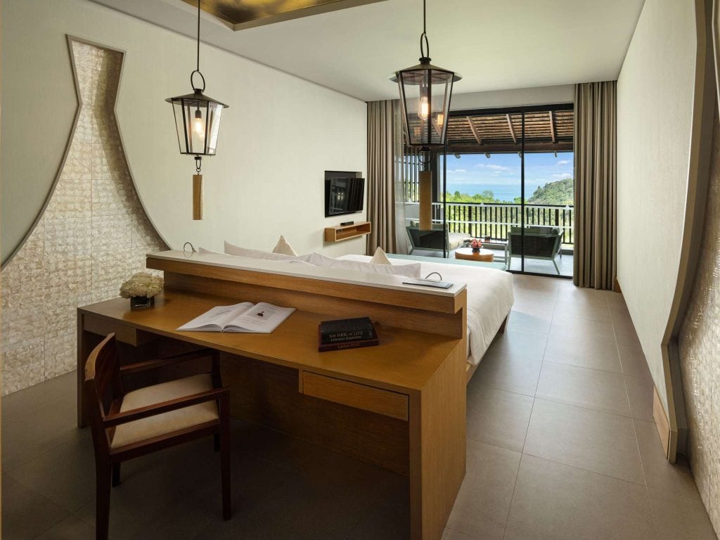 Modern hotel room with a double bed, wooden desk and chair, pendant lights, flat-screen TV, and sliding doors leading to a balcony with outdoor seating and a scenic view of greenery and mountains.