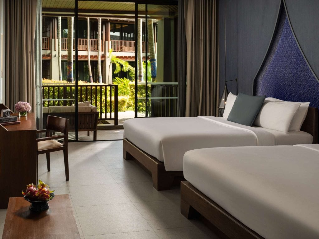 Modern hotel room with two double beds, a wooden desk and chair, and a small balcony outside with greenery and another building visible through glass doors. A bowl of fruit and flowers are on the desk.