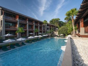 A modern hotel features a large outdoor swimming pool surrounded by sun loungers and umbrellas, with palm trees and balconies overlooking the pool under a bright blue sky.