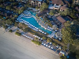 Aerial view of a beachfront resort with a large pool surrounded by blue and white umbrellas, lounge chairs, palm trees, and buildings near a sandy beach.