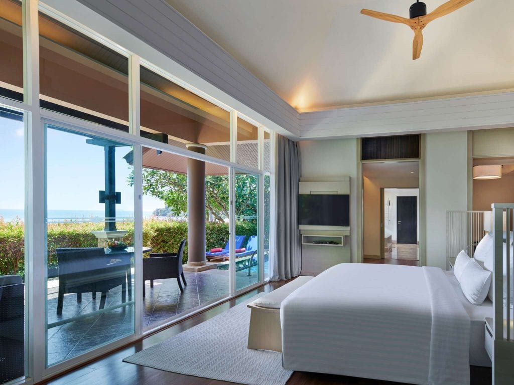 Modern hotel room with a large bed, TV, and floor-to-ceiling windows overlooking a patio with outdoor seating, greenery, and a pool, with a view of the ocean in the background.