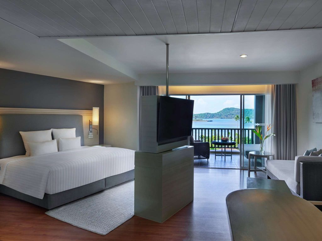 Modern hotel room with a large bed, flat-screen TV, seating area, and a balcony overlooking water and distant hills. The room has neutral decor and large windows letting in natural light.