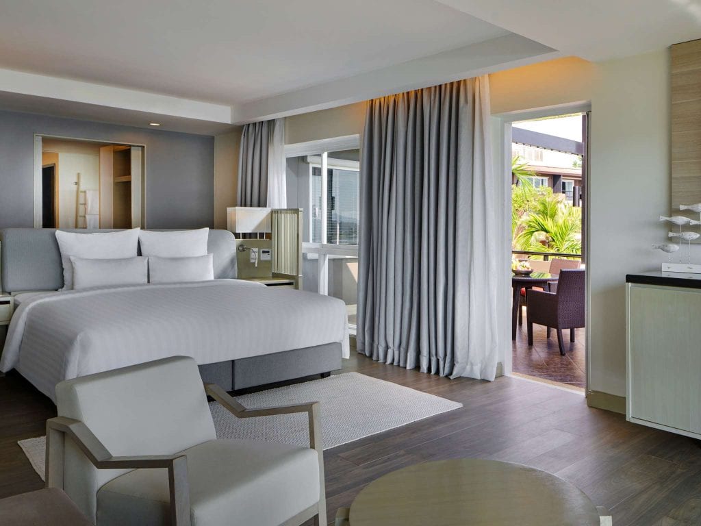 Modern hotel room with a large bed, white bedding, armchair, small table, and open curtains leading to a balcony with patio furniture and greenery outside. Warm, neutral tones create a welcoming atmosphere.