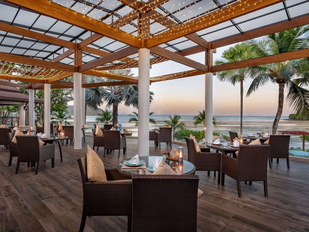 Outdoor restaurant with wicker chairs and tables set for dining, under a pergola with string lights. The area overlooks a beach with palm trees and calm ocean waves at sunset.