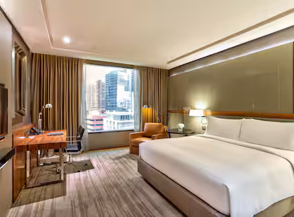 A modern hotel room with a large bed, armchair, work desk, lamps, and a window offering a city view with tall buildings. The room has neutral tones and contemporary furnishings.