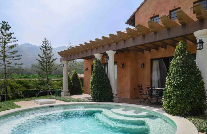 A villa with terracotta walls and a covered patio, overlooks a round pool. There are trimmed shrubs, tall trees, outdoor seating, and a scenic mountain view in the background under a clear sky.