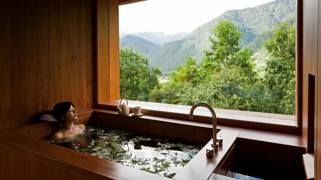 A person relaxes in a wooden soaking tub filled with water and leaves, next to large windows overlooking lush green trees and distant mountains under a bright, cloudy sky.