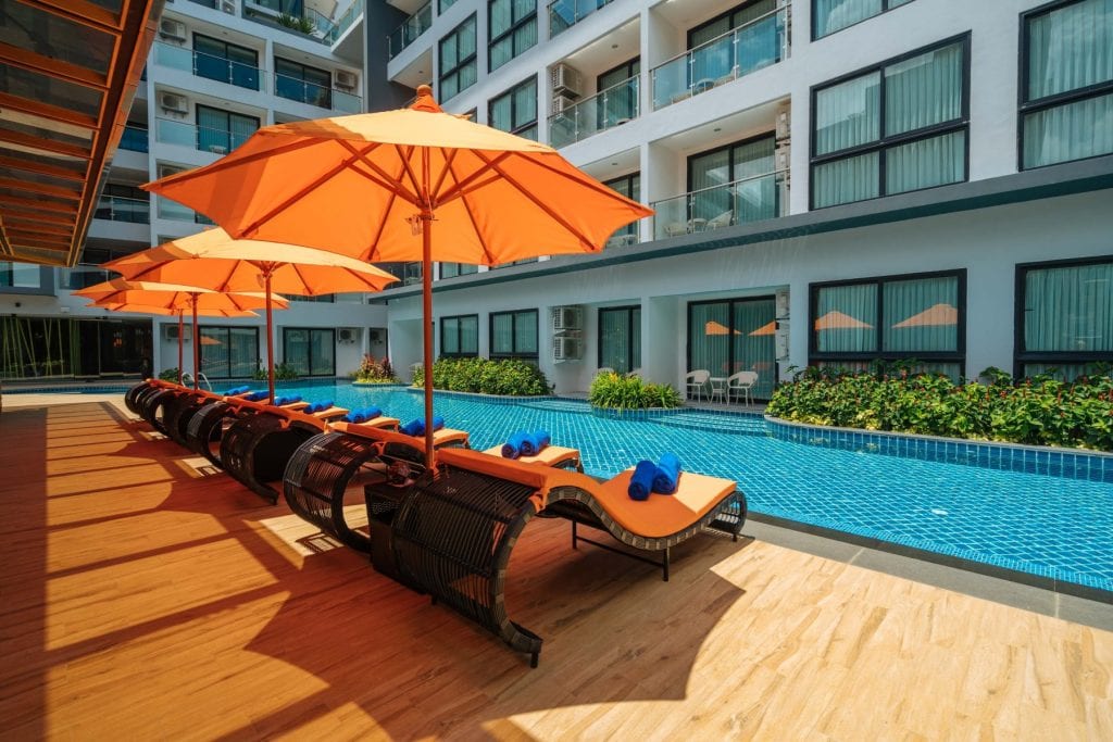 Poolside area at a modern hotel with orange lounge chairs and umbrellas, wooden deck, and a blue-tiled swimming pool. Several windows and balconies are visible on the building in the background.