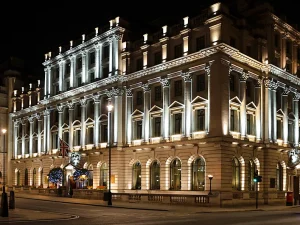 A grand, historic building is illuminated at night with elegant white lights highlighting its columns and ornate architecture. A Union Jack flag flies near the entrance, and some trees with blue lights are visible outside.