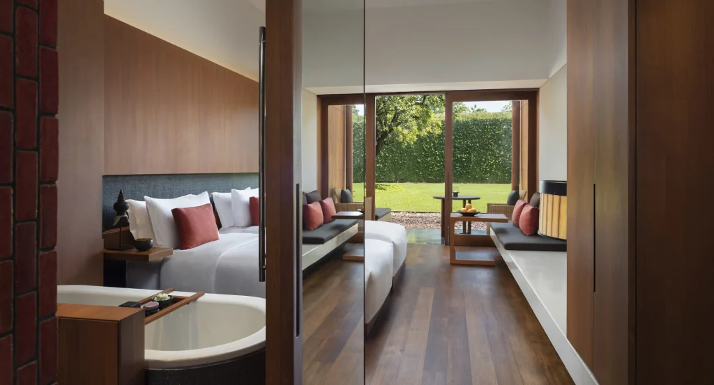 A modern hotel room with wooden floors, a bed with white linens and red pillows, a freestanding bathtub, a sitting area with a table, and large windows overlooking a green garden.