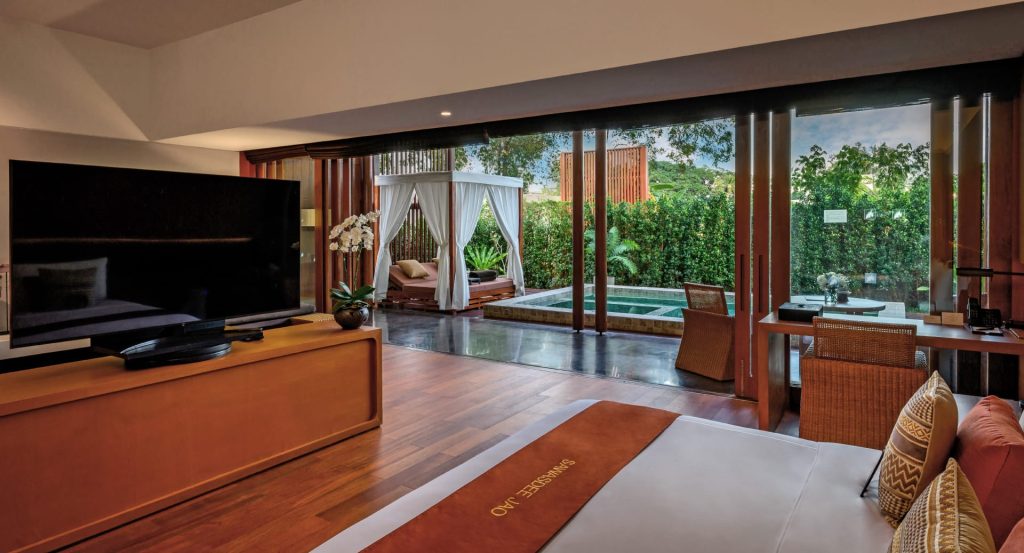 A luxurious hotel room with a large bed, a flat-screen TV, a work desk, and sliding glass doors opening to a private patio with a canopy bed, small pool, and lush greenery outside.