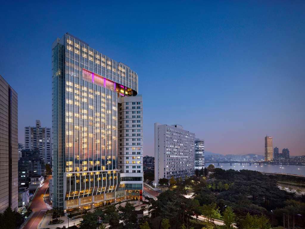 A modern high-rise hotel with illuminated windows stands among other buildings at dusk, overlooking a river and cityscape with trees and distant skyscrapers under a clear evening sky.