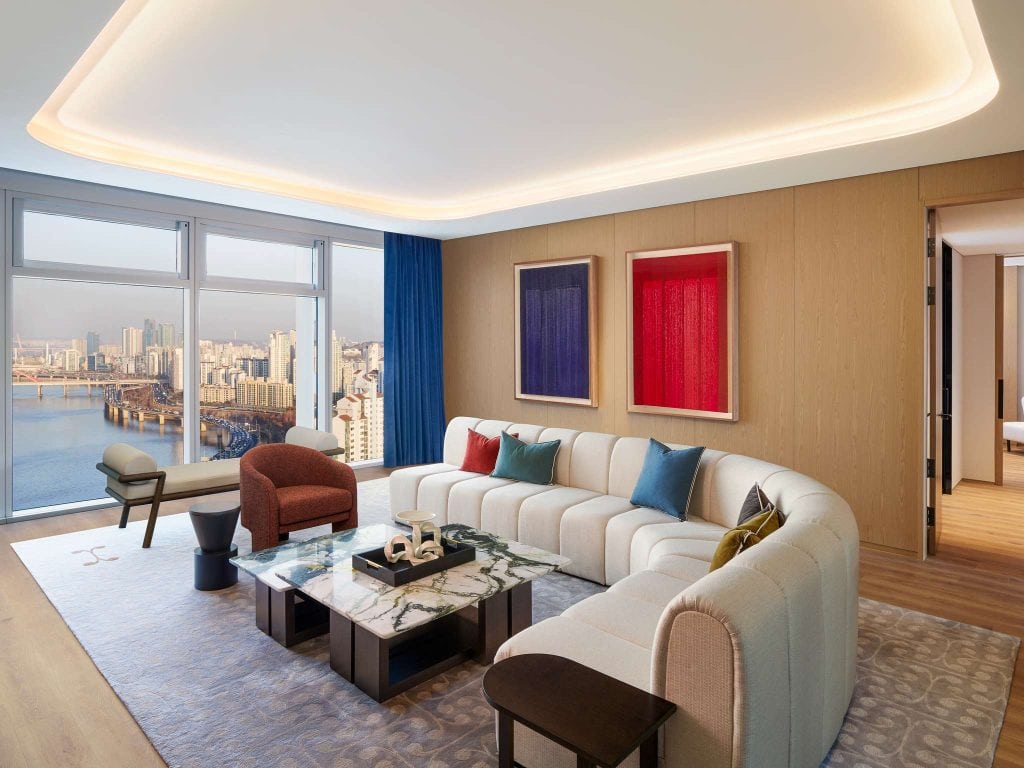 A modern living room with a large curved white sofa, colorful cushions, marble coffee tables, and contemporary art on the wall. Floor-to-ceiling windows reveal a city skyline and river view. A bedroom is visible through an open doorway.