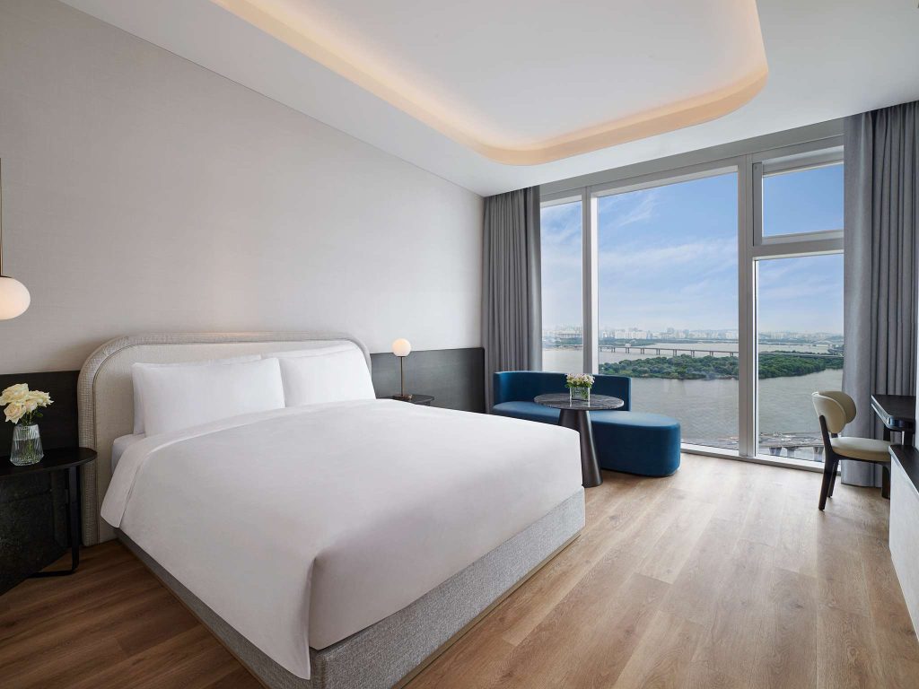 Modern hotel room with a large bed, wooden floor, bedside tables with lamps, a blue sofa by floor-to-ceiling windows, and a scenic river view outside. There is also a desk and chair near the window.