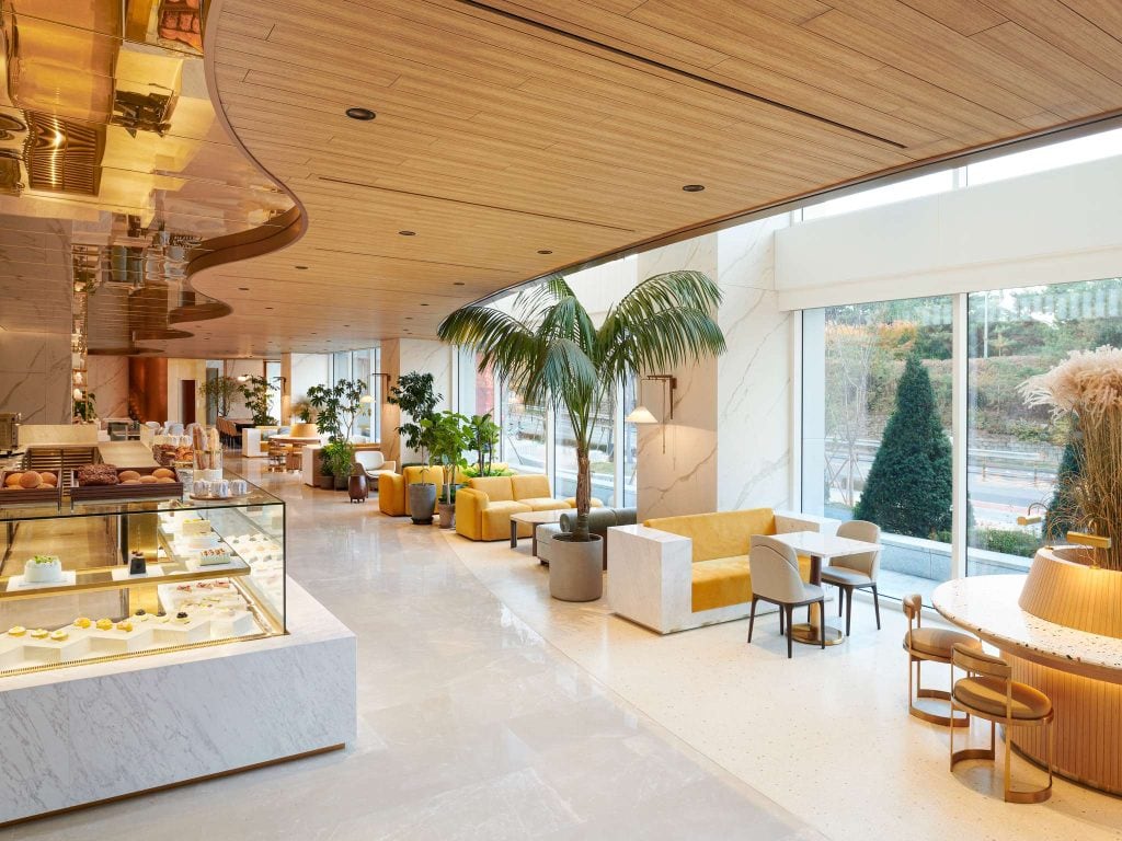 A modern, bright café features marble floors, yellow sofas, wooden ceiling panels, and large windows with lush plants and a dessert display case in the foreground. Natural light fills the inviting space.
