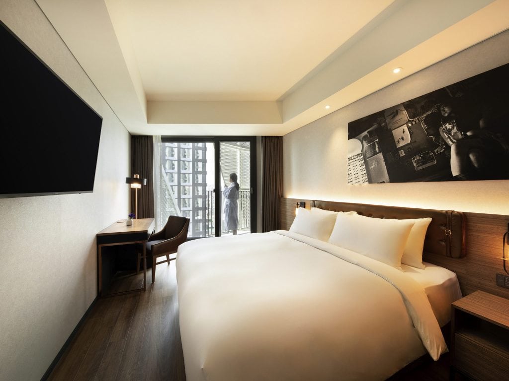 A modern hotel room with a large bed, desk, chair, wall-mounted TV, and a black-and-white photo above the headboard. A person stands on the balcony, looking outside through the glass doors.