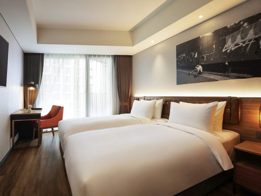 A modern hotel room with two neatly made twin beds, a wooden floor, a desk with an orange chair, floor-to-ceiling window with sheer curtains, and a large black-and-white photo on the wall above the beds.