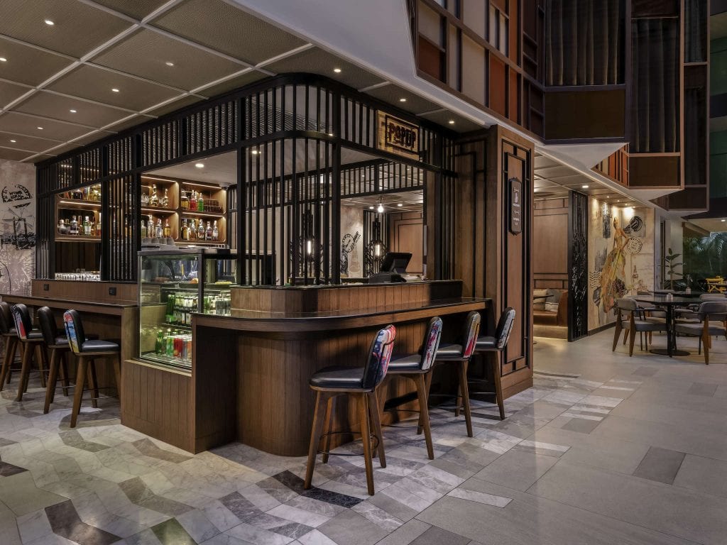 Modern hotel bar with wooden counters, high-backed barstools, and shelves stocked with bottles. The area is well-lit, with seating nearby and stylish décor featuring geometric patterns and wall art.