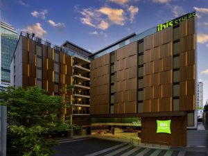 Modern, multi-story Ibis Styles hotel with a wooden-panel facade, large windows, and green signage. The building is surrounded by trees and set against a blue sky with clouds at sunset.