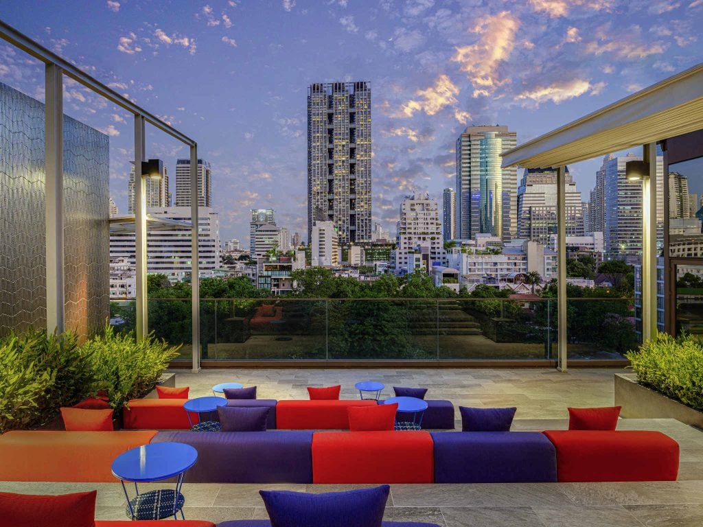 A rooftop lounge with colorful red, blue, and purple seating overlooks a city skyline with tall buildings and lush green trees under a vibrant, cloud-streaked evening sky.
