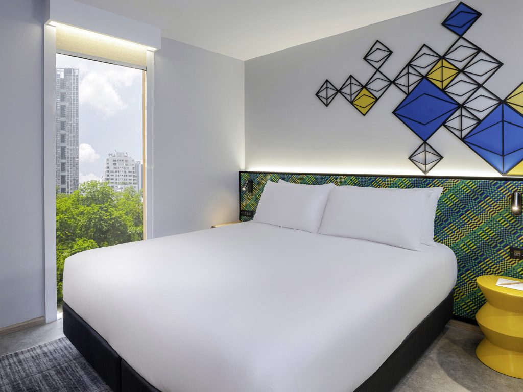 A modern hotel room with a large neatly made bed, colorful geometric wall art, a yellow nightstand with a lamp, and a floor-to-ceiling window showing city buildings and green trees outside.