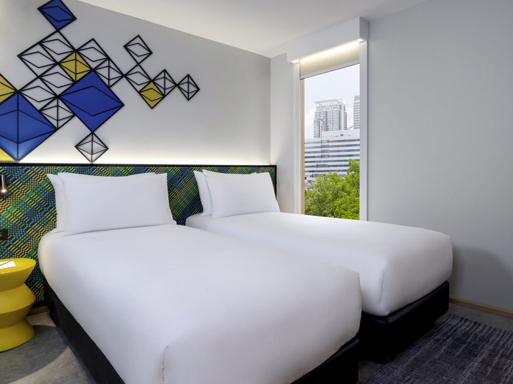 A modern hotel room with two neatly made single beds, a yellow side table, geometric wall art above the headboard, and a large window showing city buildings and greenery outside.