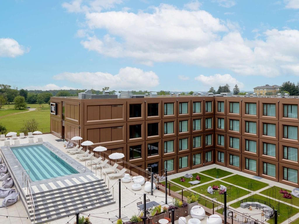 A modern hotel with a brown facade surrounds a courtyard featuring a rectangular pool with lounge chairs and white umbrellas, and a green lawn area with scattered seating, set under a blue sky with clouds.