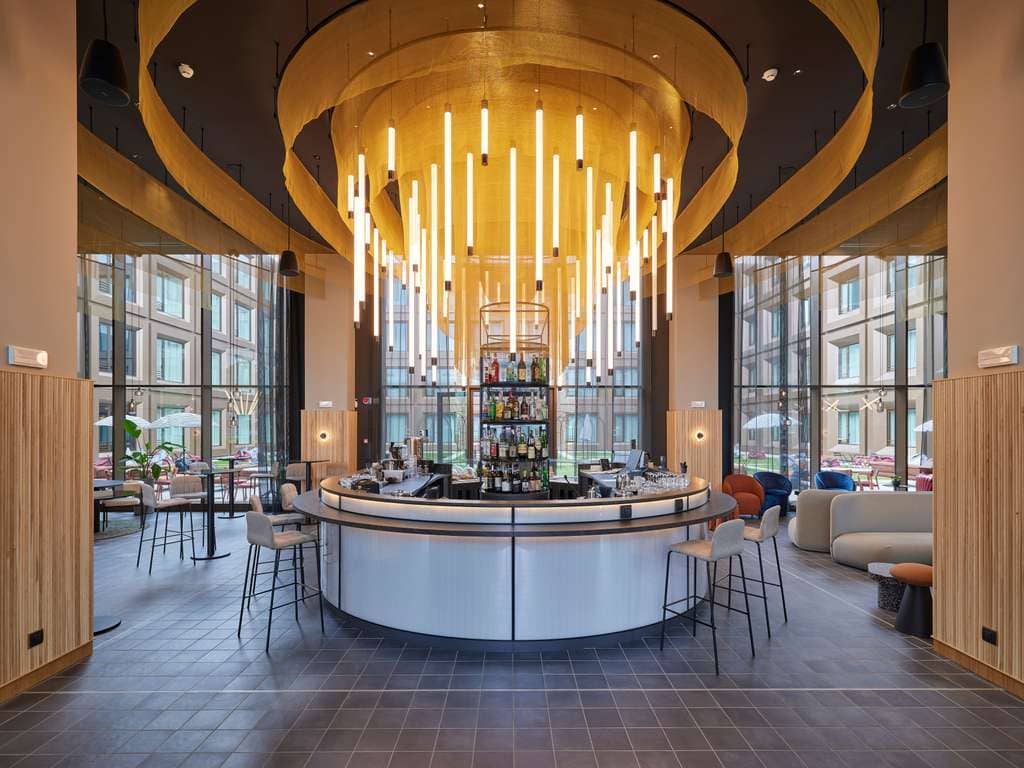 A modern circular bar with high chairs, surrounded by large windows. Dramatic, vertical pendant lights hang from a gold spiral ceiling feature. The setting is stylish and bright, with seating and patio views outside.