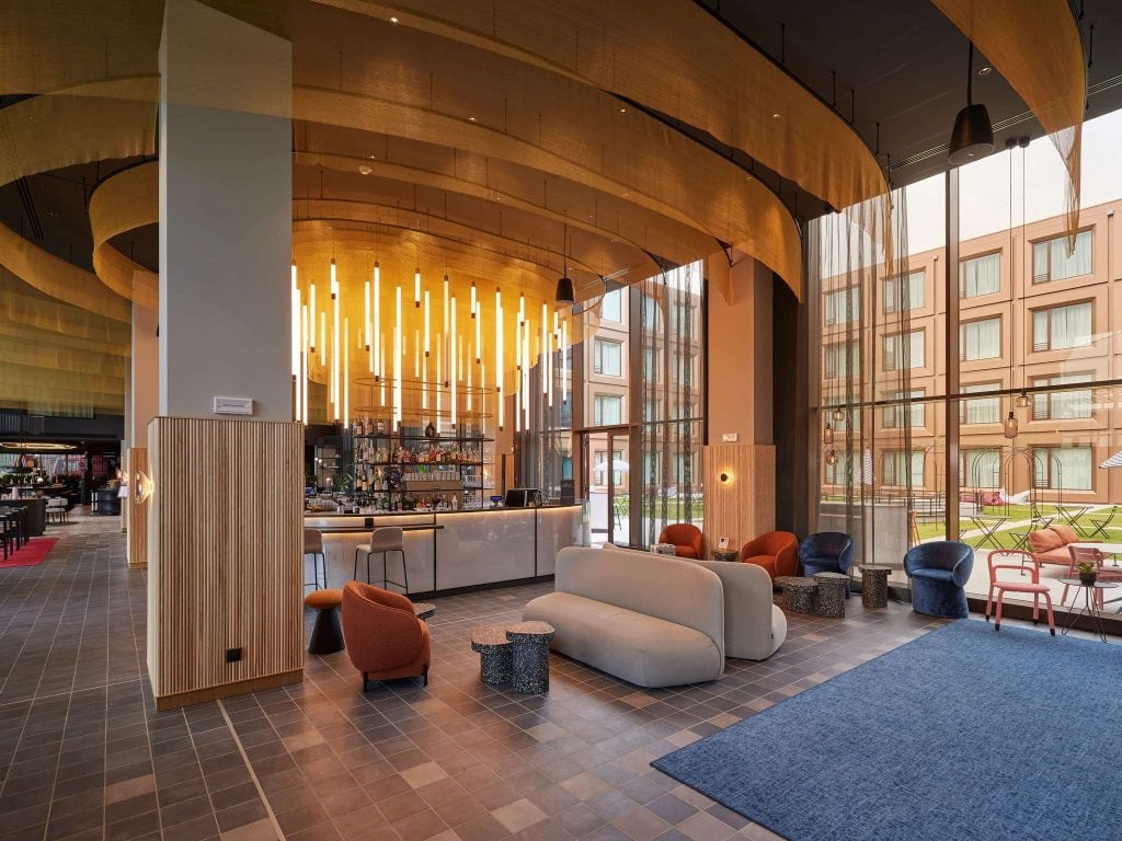 Modern hotel lobby with stylish seating, a central white bar, warm overhead lighting fixtures, large windows, and a view of the hotel’s outdoor area and rooms. Contemporary décor in neutral and warm tones.