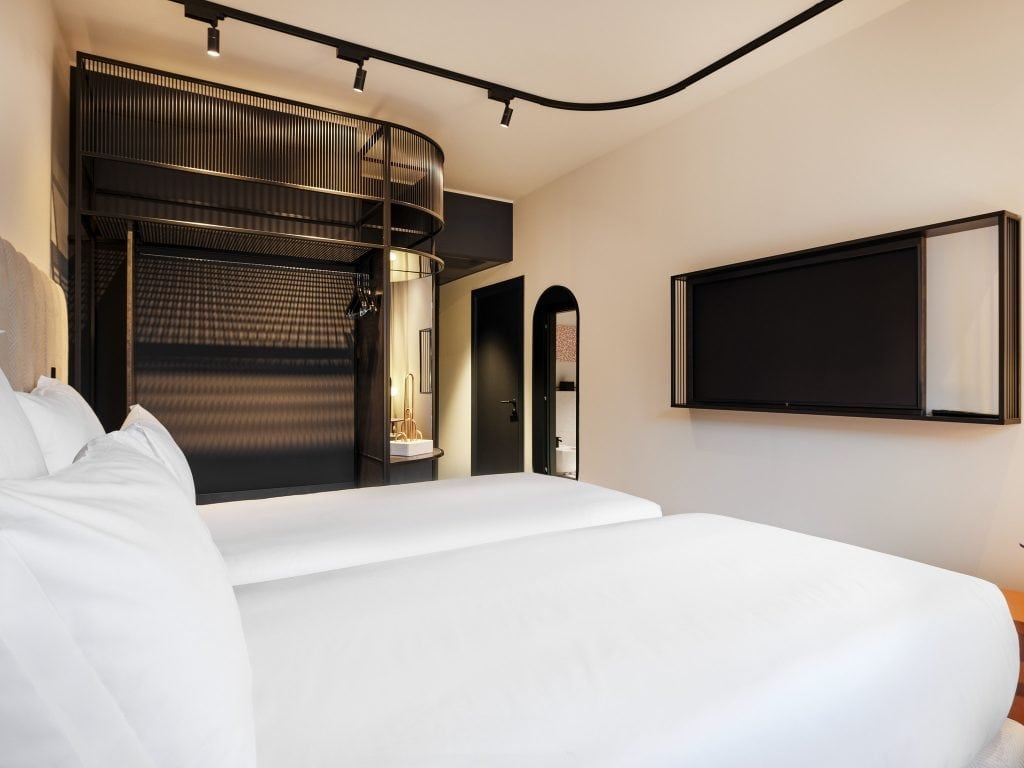 Minimalist hotel room with two neatly made beds, a large wall-mounted flat-screen TV, an open black wardrobe, and a modern bathroom sink area with round mirror in the background. The decor is sleek and contemporary.