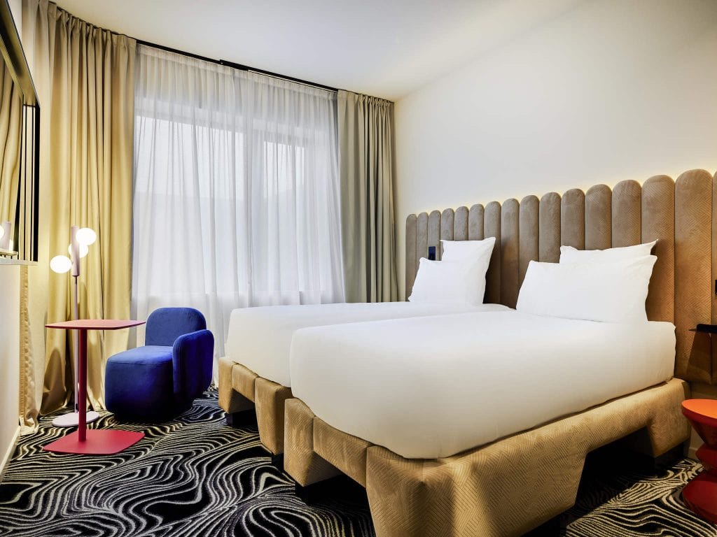 A modern hotel room with two twin beds, white bedding, a beige padded headboard, a unique black-and-white patterned carpet, a blue chair, a red side table, and a lamp near a large window with sheer curtains.
