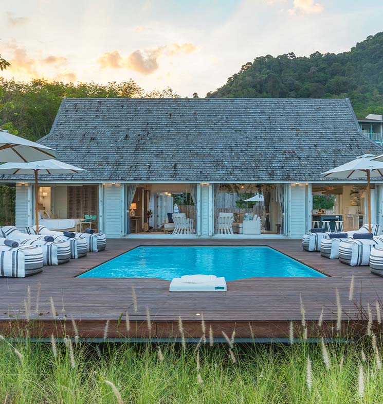 A luxurious villa with a swimming pool, striped lounge chairs, and umbrellas sits in front of a wooden house with large windows, surrounded by greenery and hills at sunset.