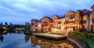 Row of Mediterranean-style townhouses with warm lights reflected on a calm lake at dusk, featuring balconies and a central patio area surrounded by greenery and trees.