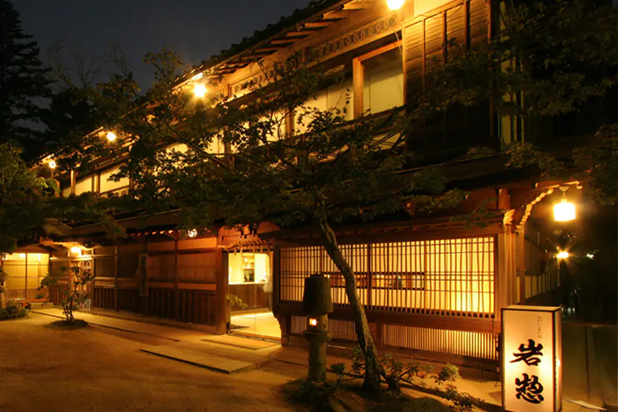 A traditional Japanese inn with wooden architecture is warmly illuminated at night. Soft yellow lights glow from inside and outside, highlighting wooden details and sliding doors. A lit sign stands near the entrance.