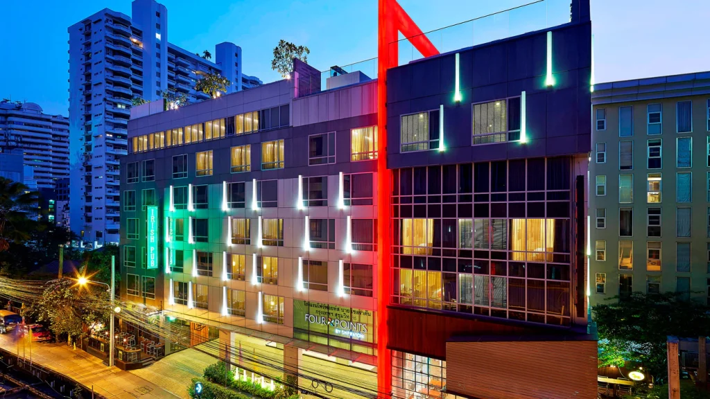 A modern hotel building at dusk with illuminated windows and red, green, and white lights on its facade. Tall buildings and city lights are visible in the background.