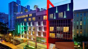 A modern hotel building at dusk with illuminated windows and red, green, and white lights on its facade. Tall buildings and city lights are visible in the background.